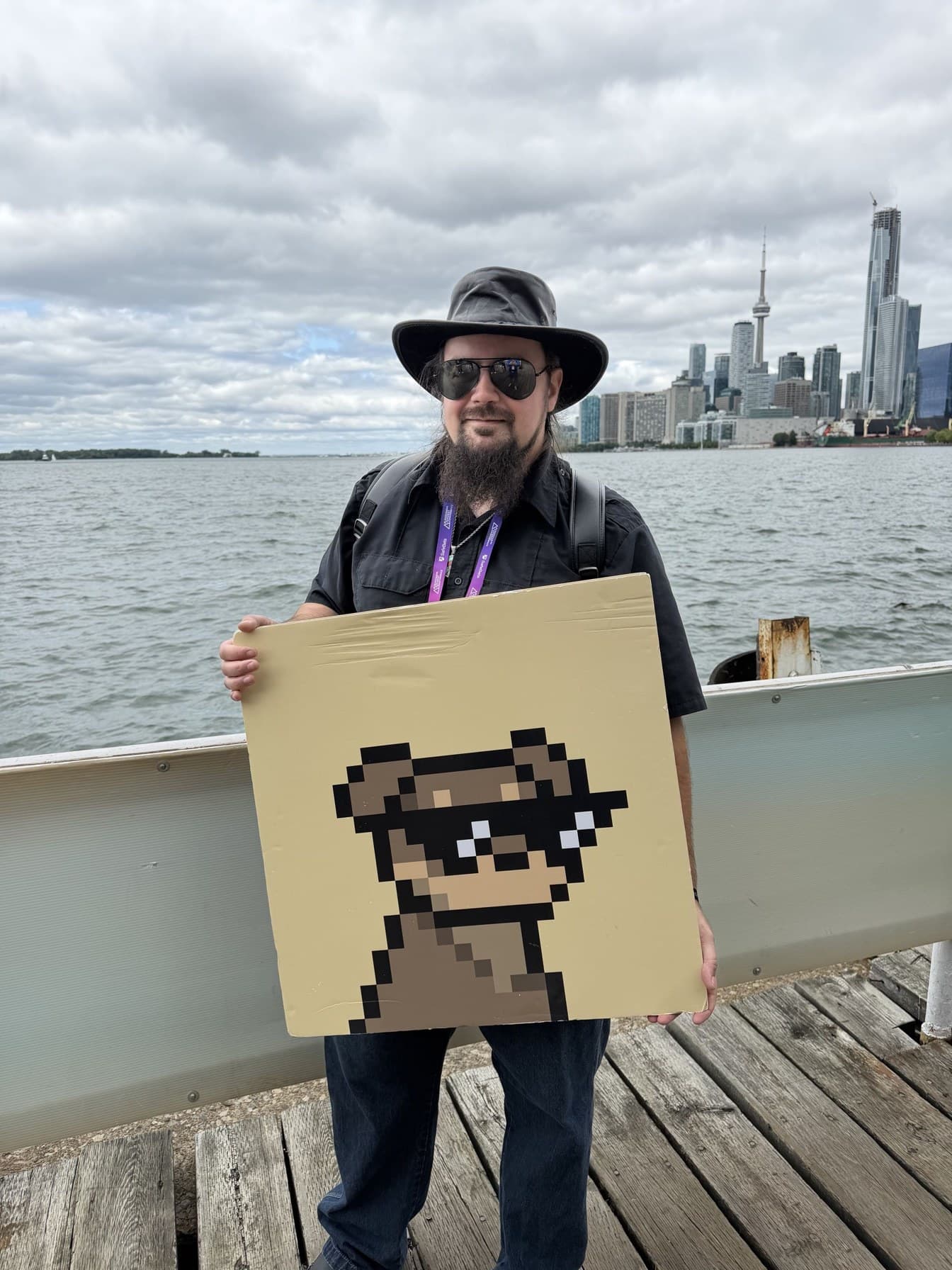 Metal (MetalXMeta) holding a handmade pixel board Metal (MetalXMeta) in a black hat and sunglasses holding a large pixel-art board on a waterfront